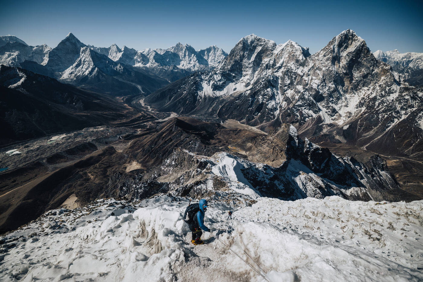 A climber ascending the snowy summit ridge of Lobuche East seen from above, with Himalayan peaks and valleys beyond