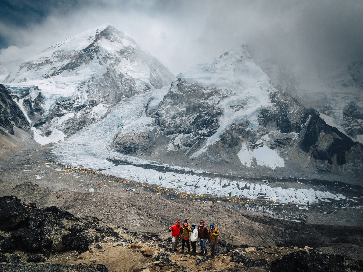 Five team members posing on a rocky hillside above Base Camp with the Khumbu Icefall and glacier behind