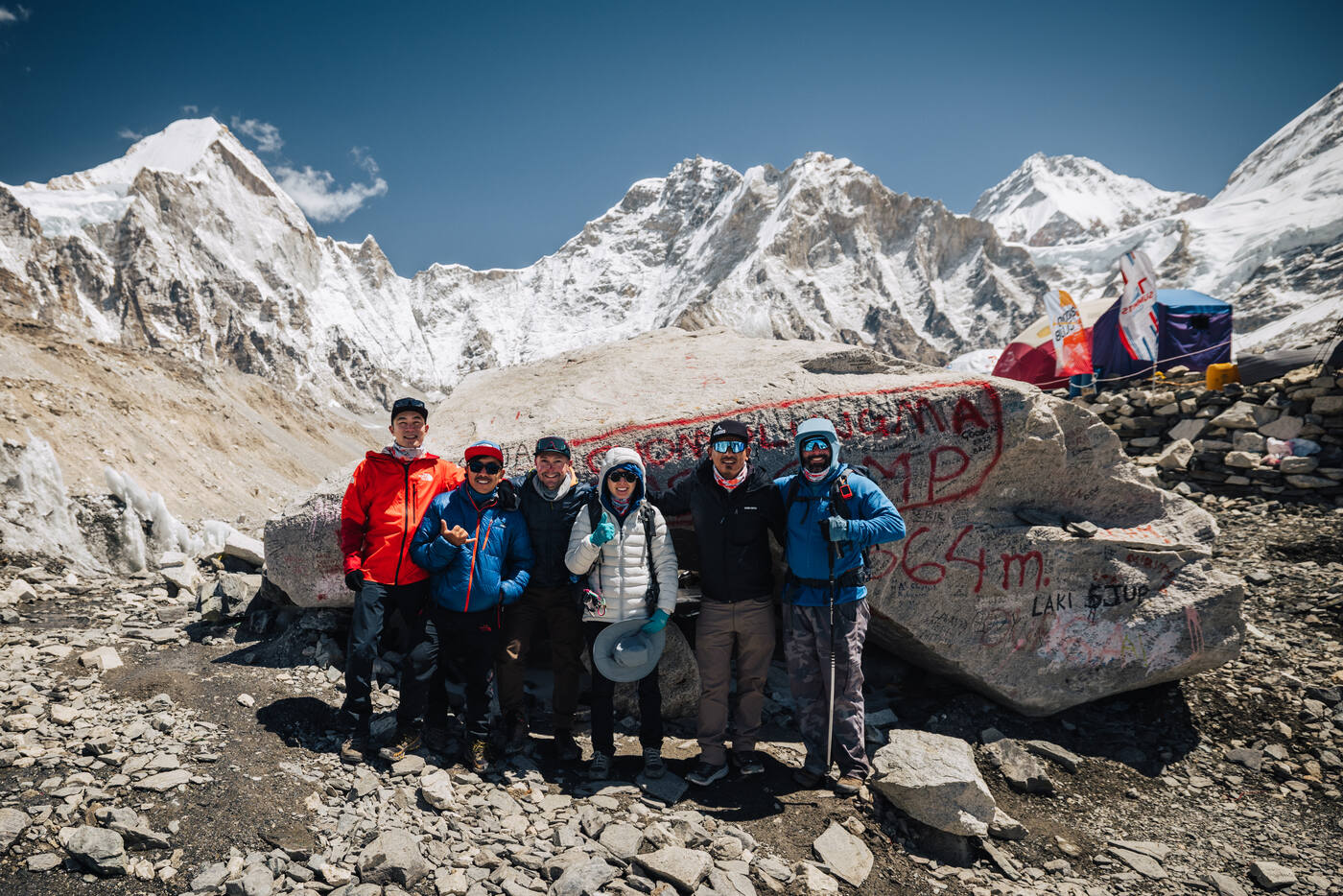 Group of six trekkers and climbers posing in front of the Everest Base Camp rock with prayer flags and peaks behind