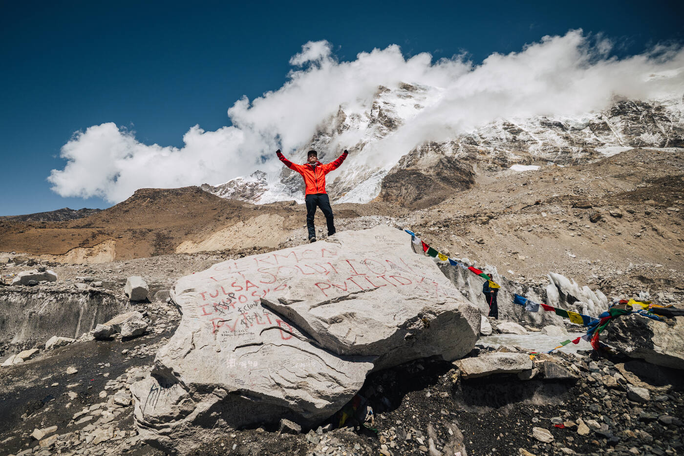 Jason in a red jacket standing on the Everest Base Camp rock with arms raised, clouds and peaks behind