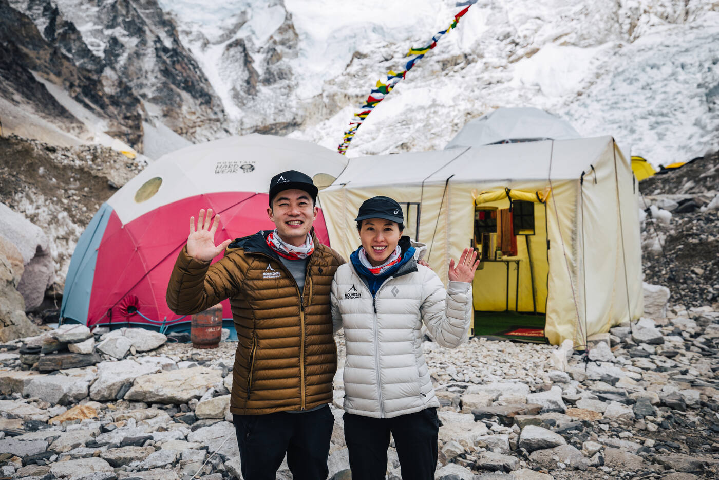 Jason and Grace waving outside the dome tent at Everest Base Camp with prayer flags overhead