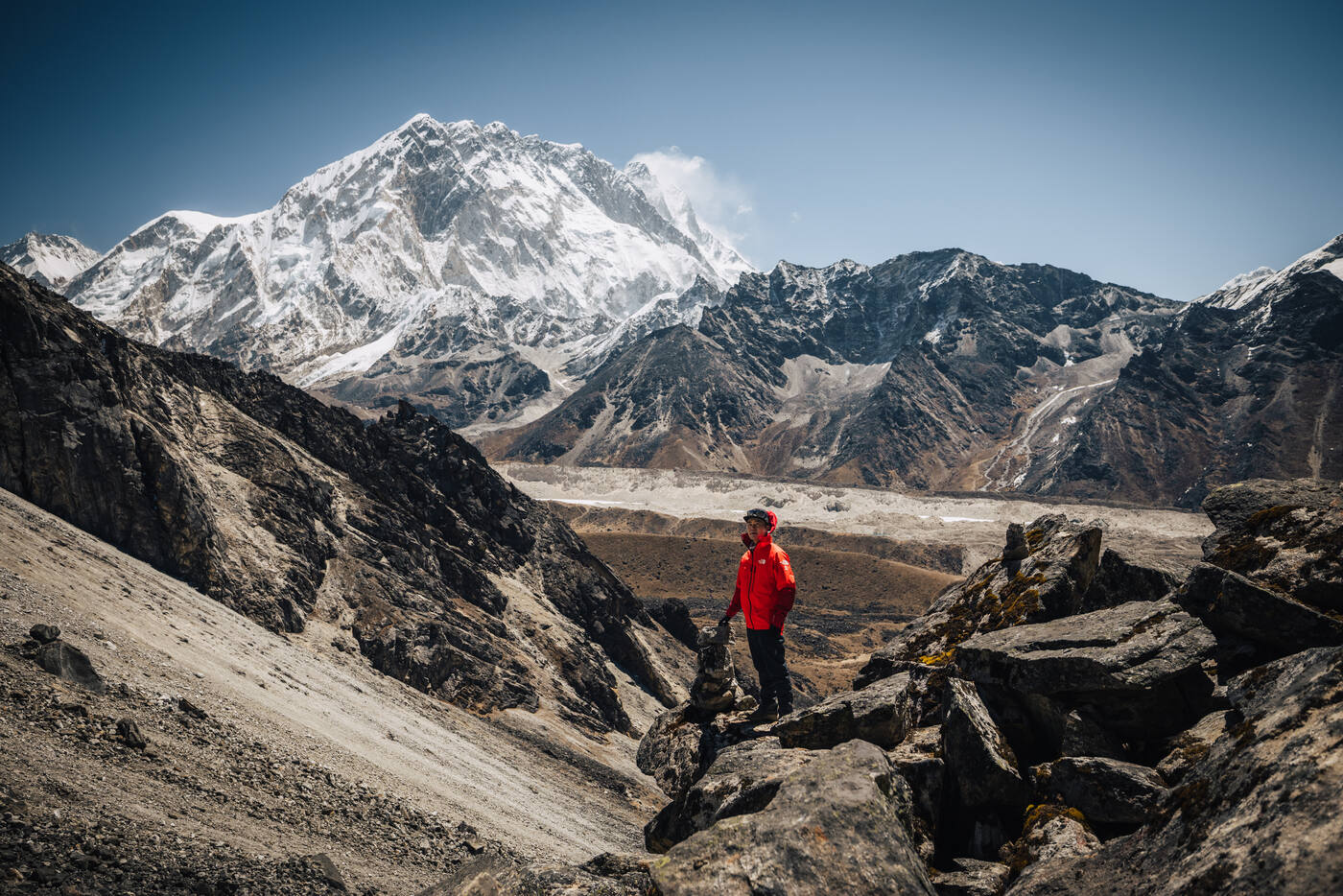 A climber in a red jacket on a rocky ridge with a large snow peak and glacier valley behind