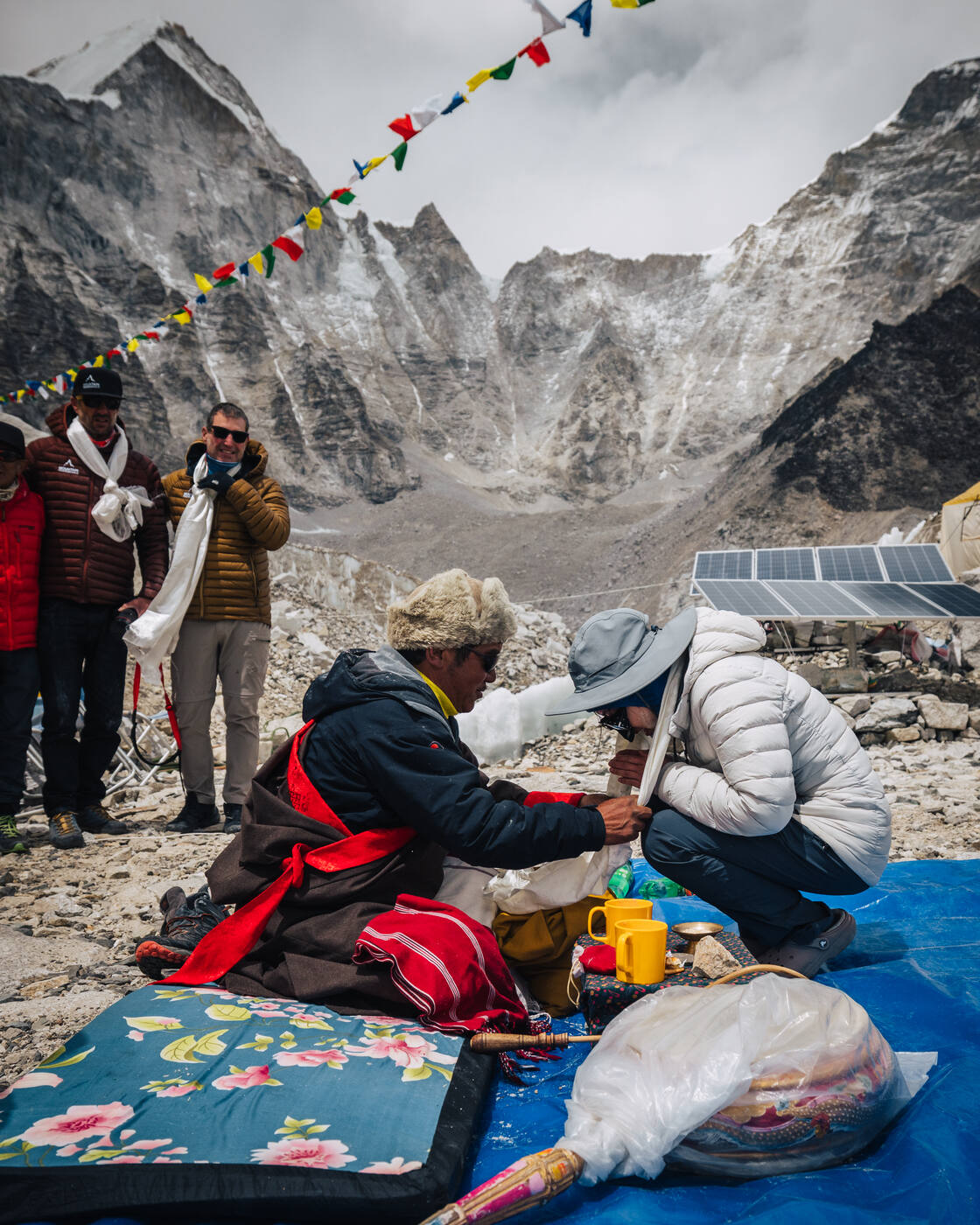 The lama blessing a kneeling climber in a white jacket at the Puja with prayer flags above
