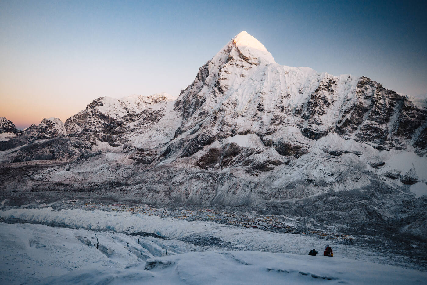 A snow pyramid peak catching first light at dawn above the glacier, with two people seated in the foreground