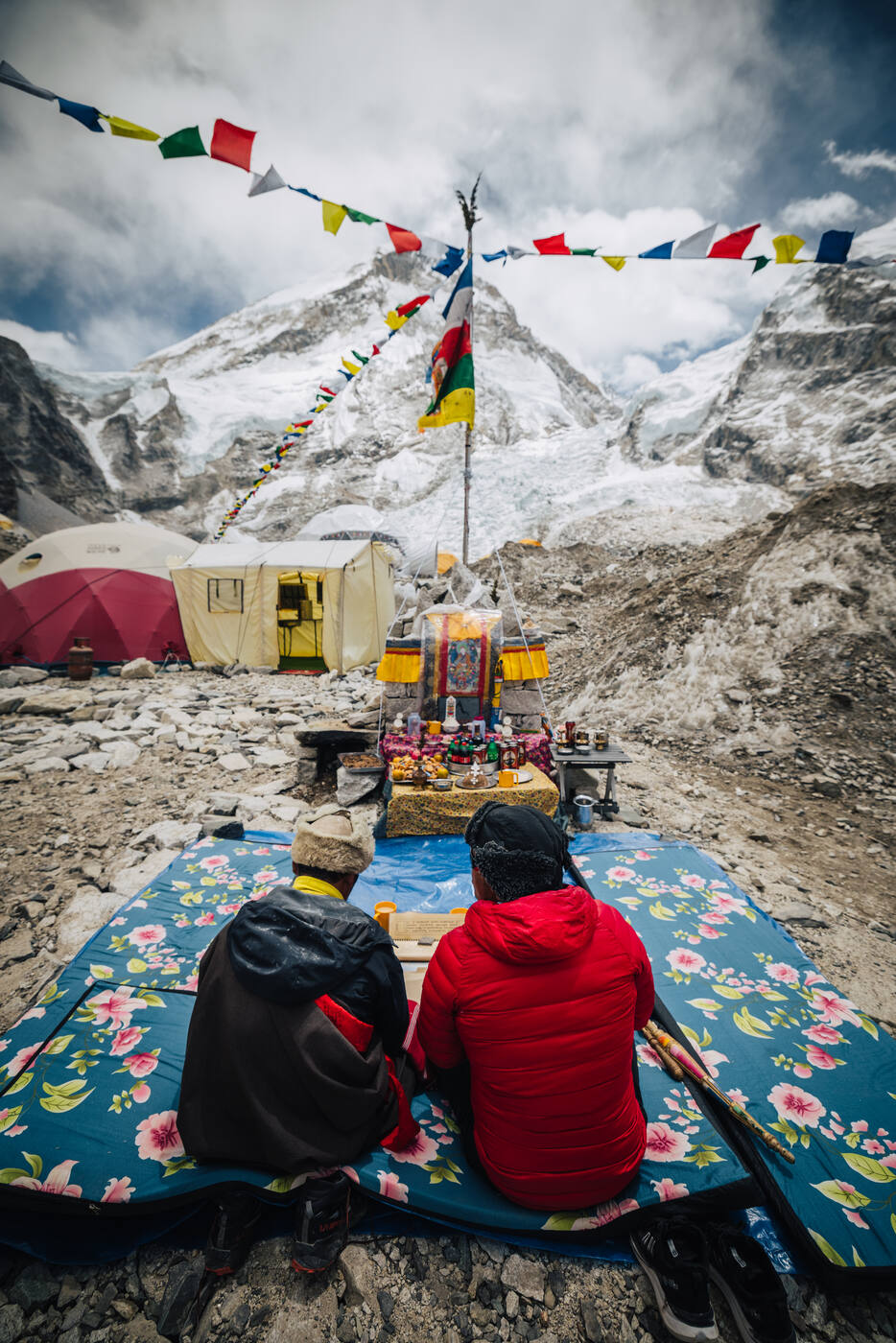 Two climbers seated before the Puja altar with prayer flags radiating overhead and snow peaks behind