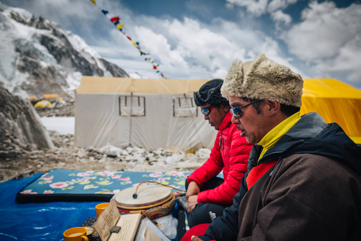 Two Sherpa team members in red jackets seated at the Puja ceremony with a drum and offerings