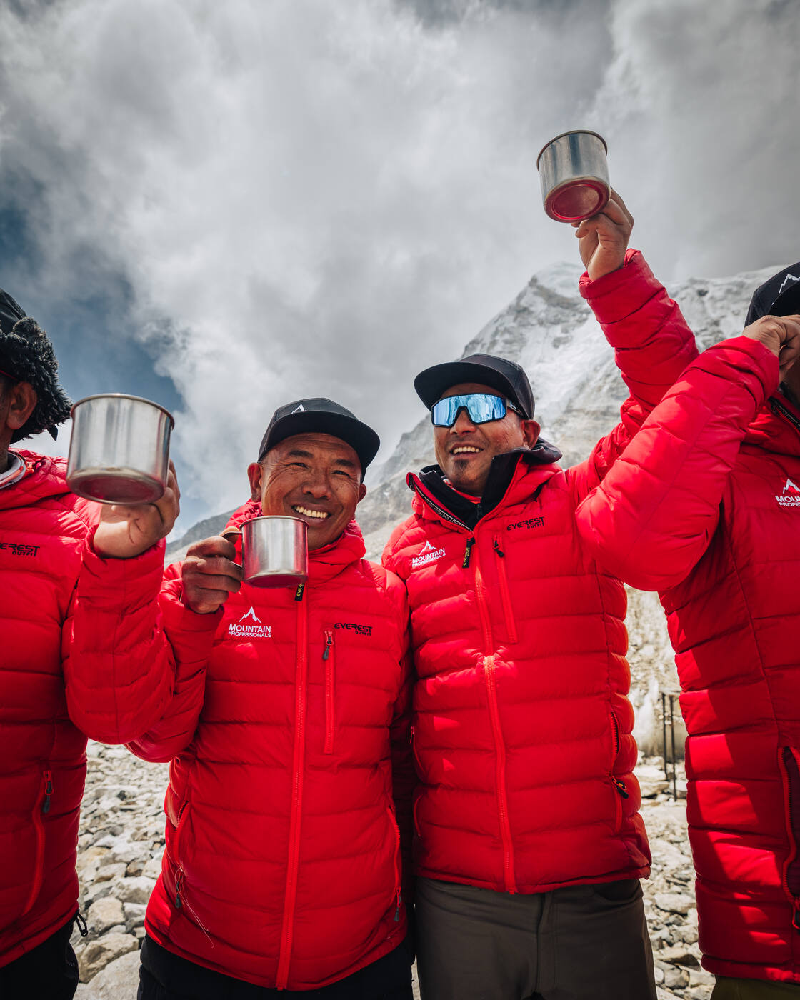 Sherpa team members in matching red jackets raising metal cups in a toast at Everest Base Camp