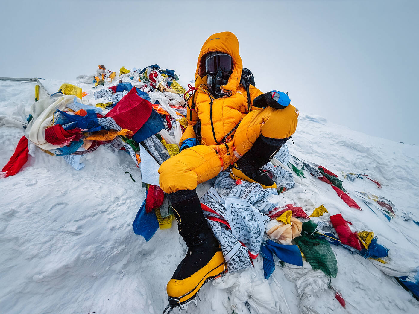 Jason sitting at the summit of Everest surrounded by prayer flags in whiteout conditions wearing yellow summit suit