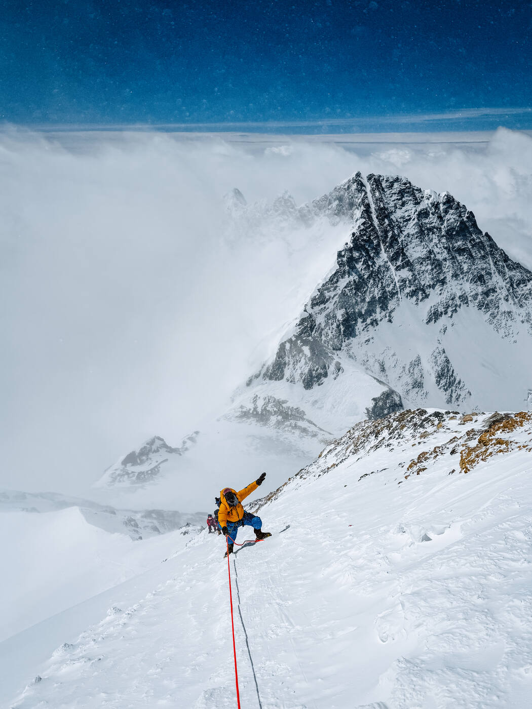 A climber in a yellow down suit on the snow slope high on Everest with a rocky peak rising above a sea of cloud behind