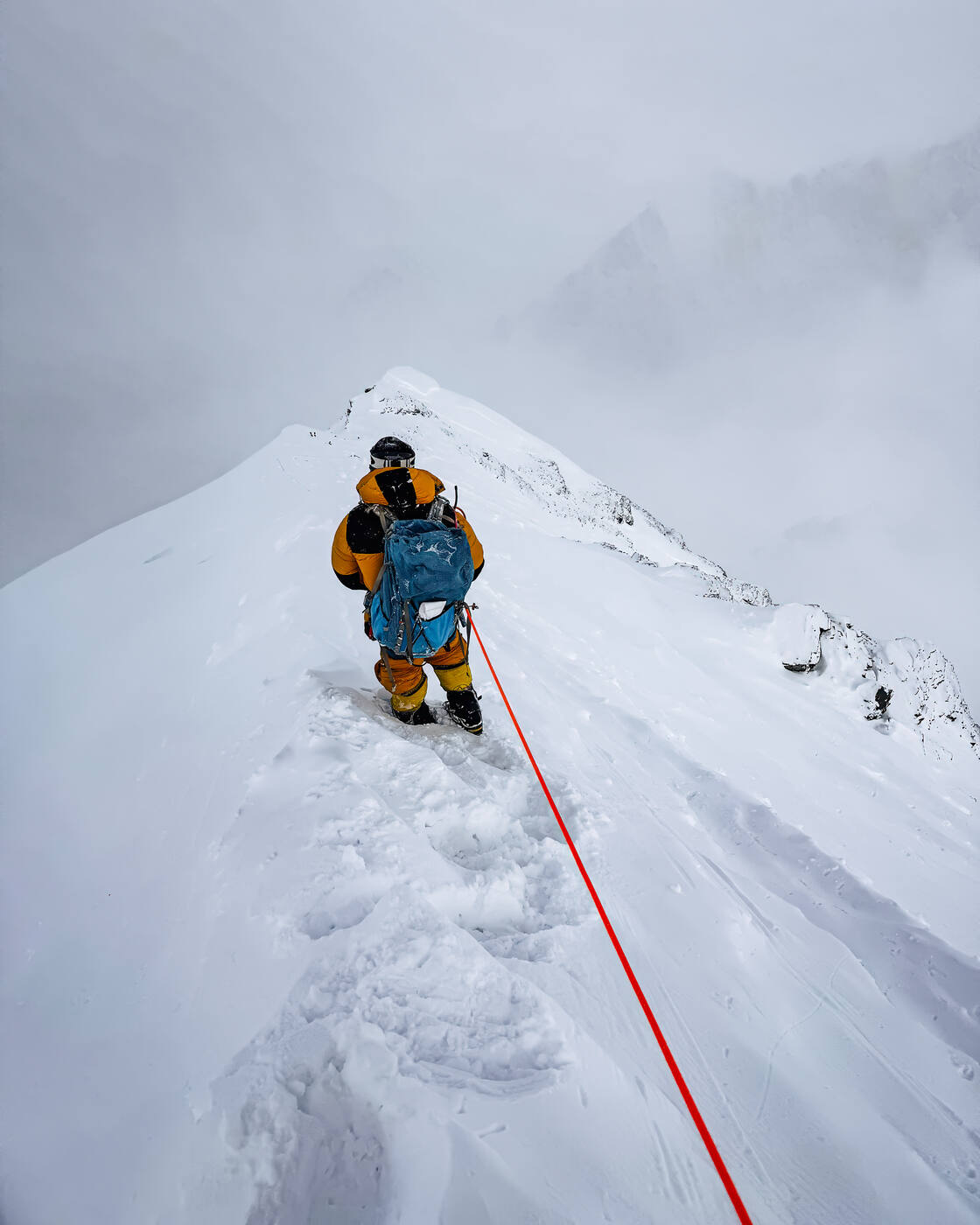 Climber in yellow summit suit walking along a narrow snow ridge on fixed rope in whiteout conditions near the summit of Everest