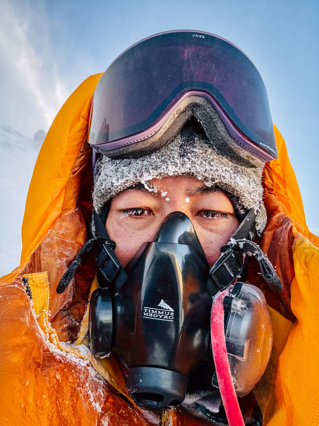 Close-up selfie of Grace wearing oxygen mask and frosted goggles in orange summit suit near the top of Everest
