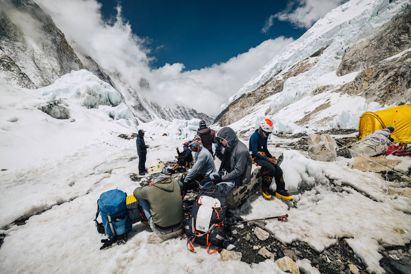 Team members resting on rocks at Camp 2 with yellow tents and mountain walls behind