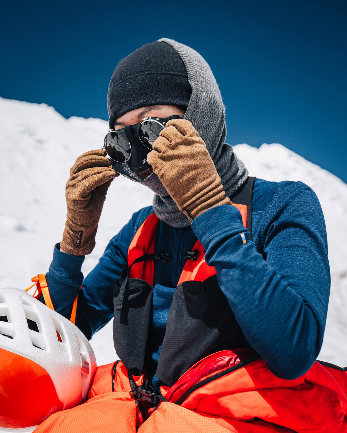 A climber in a down suit and balaclava adjusting goggles, helmet in hand
