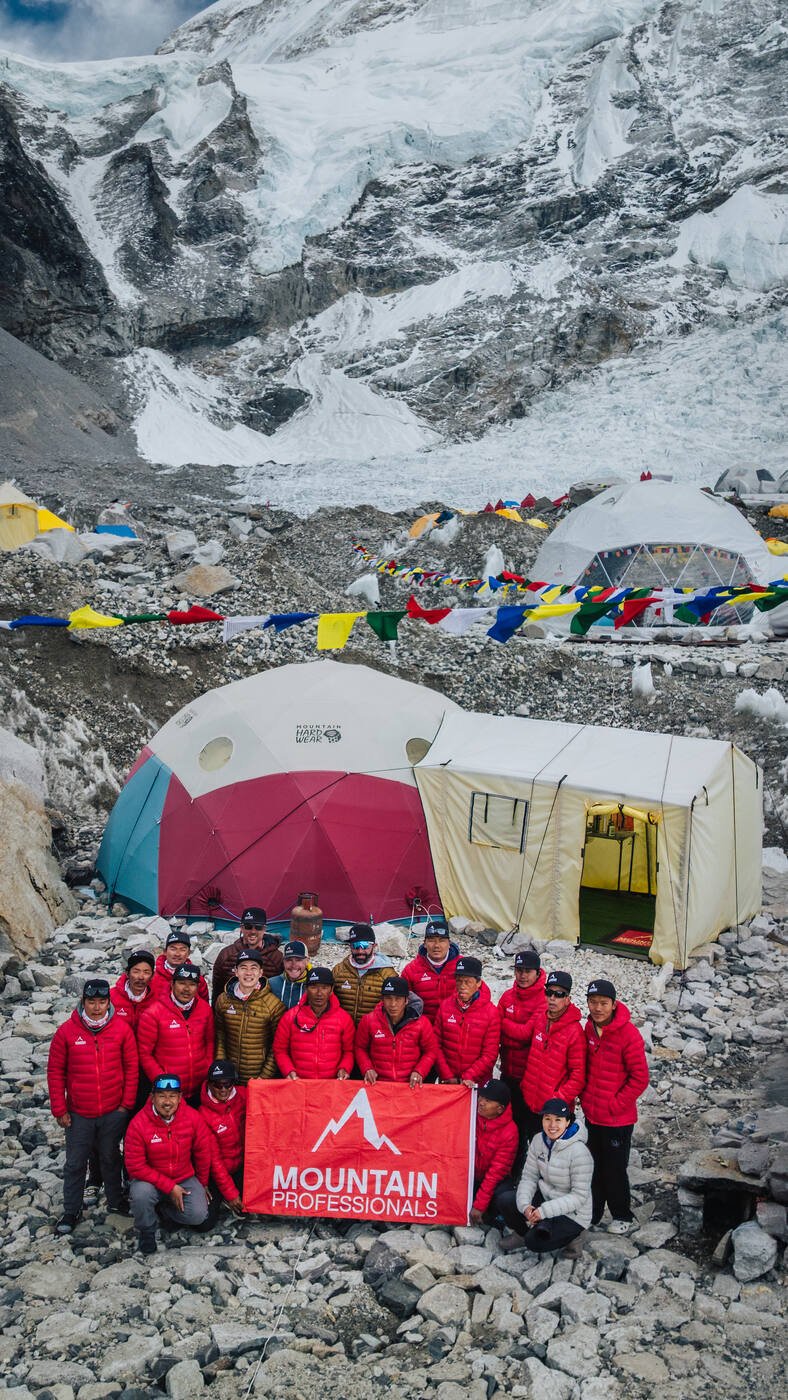 Mountain Professionals team posing with the red banner in front of the dome tents at Base Camp