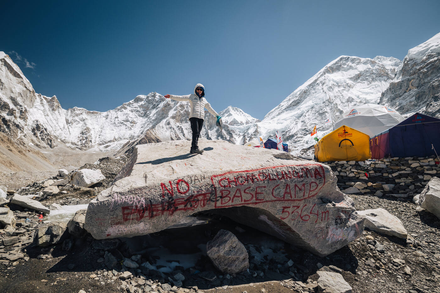 A climber standing with arms outstretched on the Everest Base Camp rock, with tents and snow peaks behind