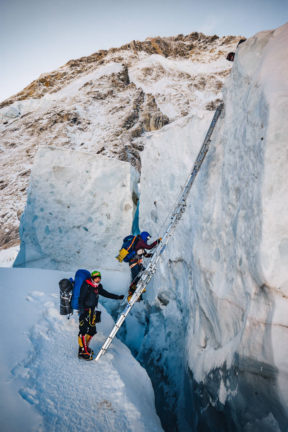 Climber ascending a vertical ladder against an ice wall deep inside a crevasse in the Khumbu Icefall