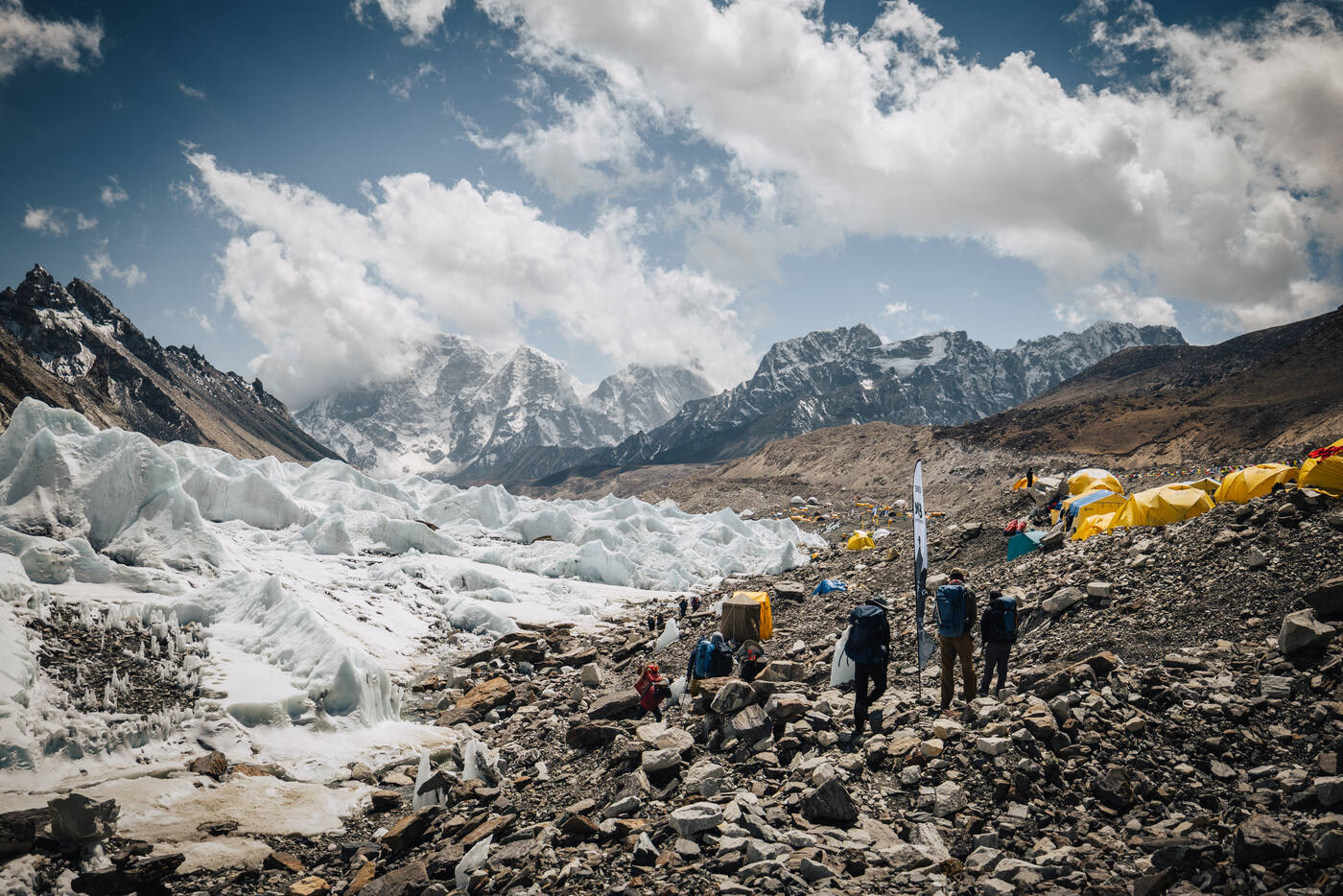 Climbers walking across the rocky glacier toward tents with peaks behind