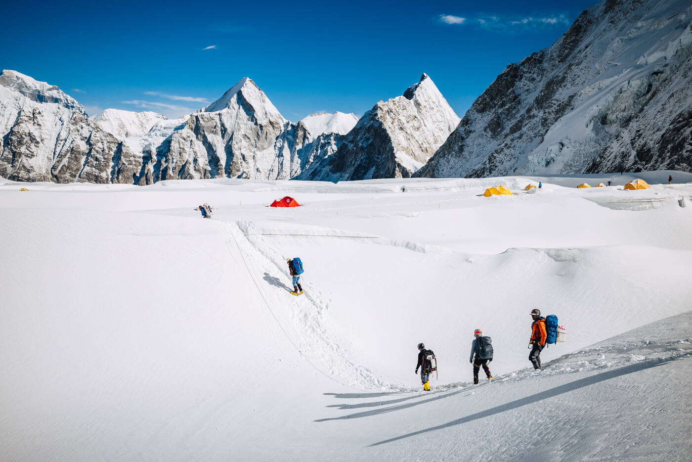 Climbers walking across flat snow in the Western Cwm near Camp 1 with dramatic peaks and colorful tents behind