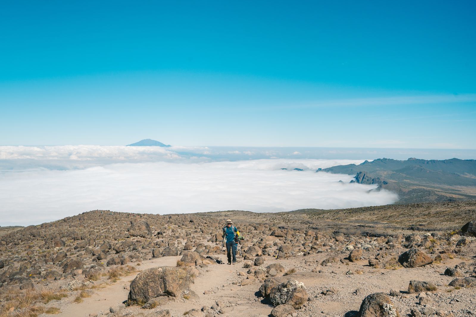 Hiker walking arid alpine terrain above a sea of clouds, Mount Meru on the horizon