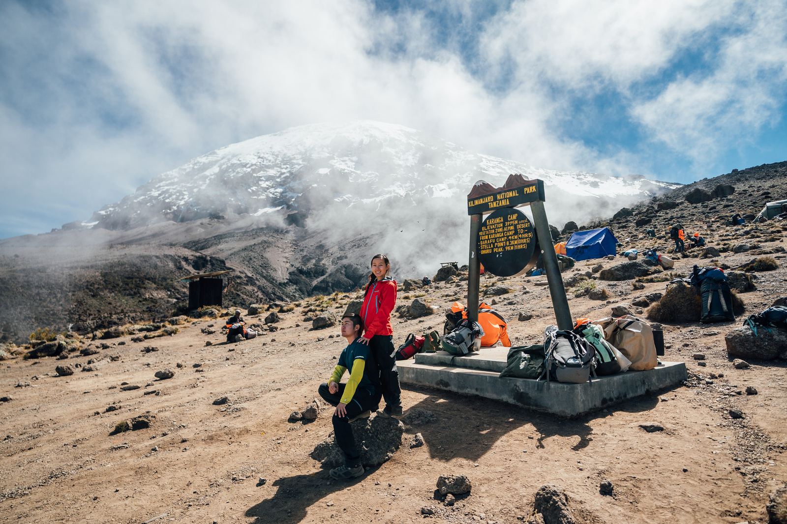 Jason and Grace resting beside a camp signboard and piled packs with Kilimanjaro's snow-covered summit wrapped in cloud behind
