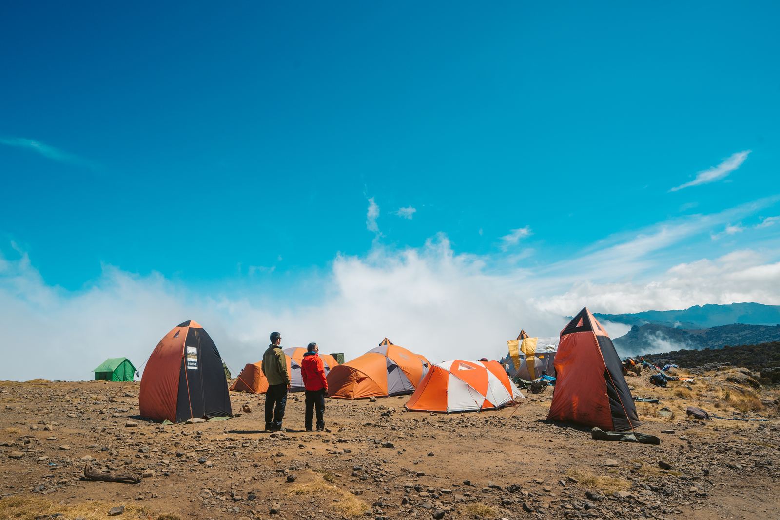 Orange and green expedition tents at Shira Camp under a bright blue sky with clouds rolling in below