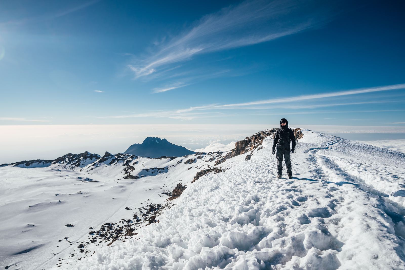 Climber walking along the snowy crater rim trail toward Uhuru Peak with Mawenzi visible