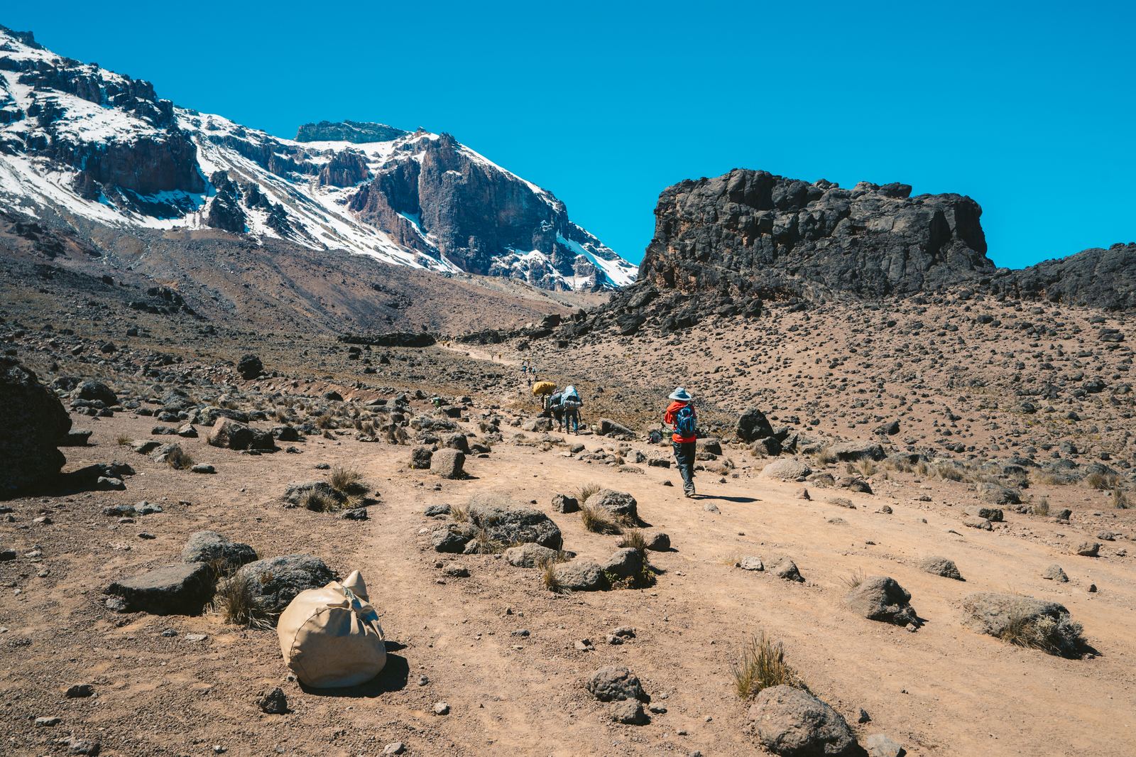 Hiker descending rocky alpine slope toward snowy Kilimanjaro peak with cloud-covered valleys below