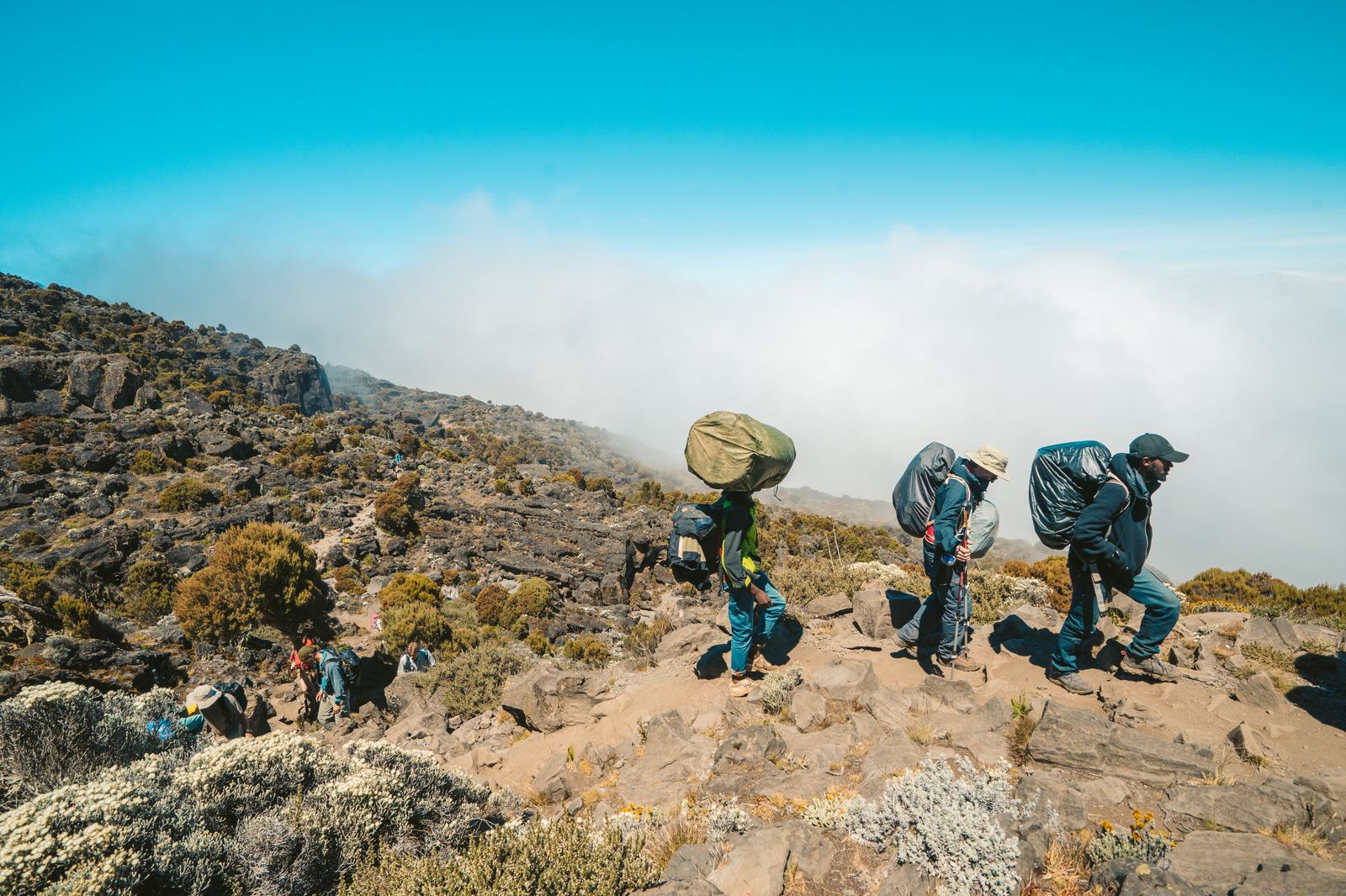 Porters carrying heavy loads along a rocky ridge above the clouds
