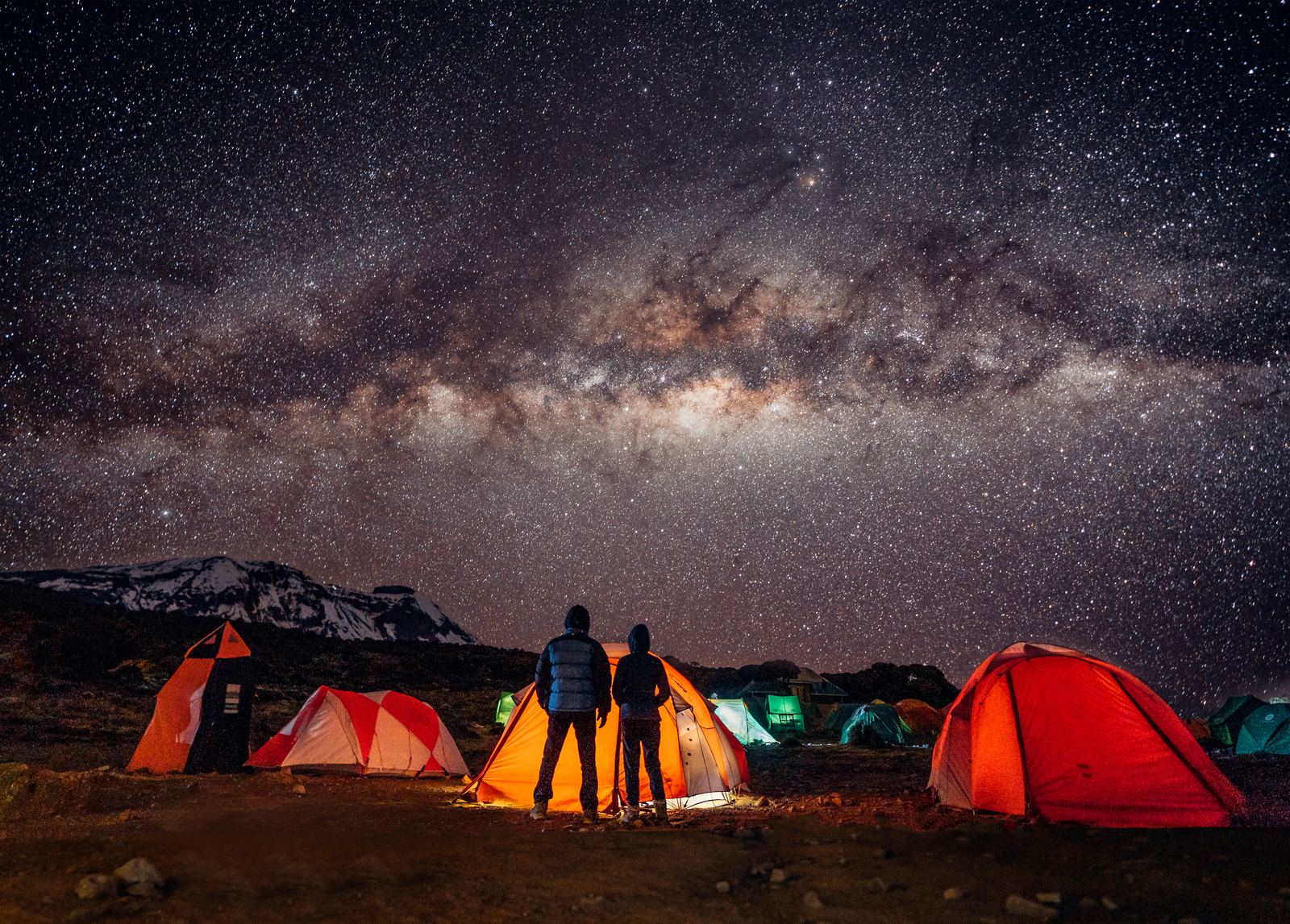 Jason and Grace silhouetted between glowing orange tents under the Milky Way on Kilimanjaro