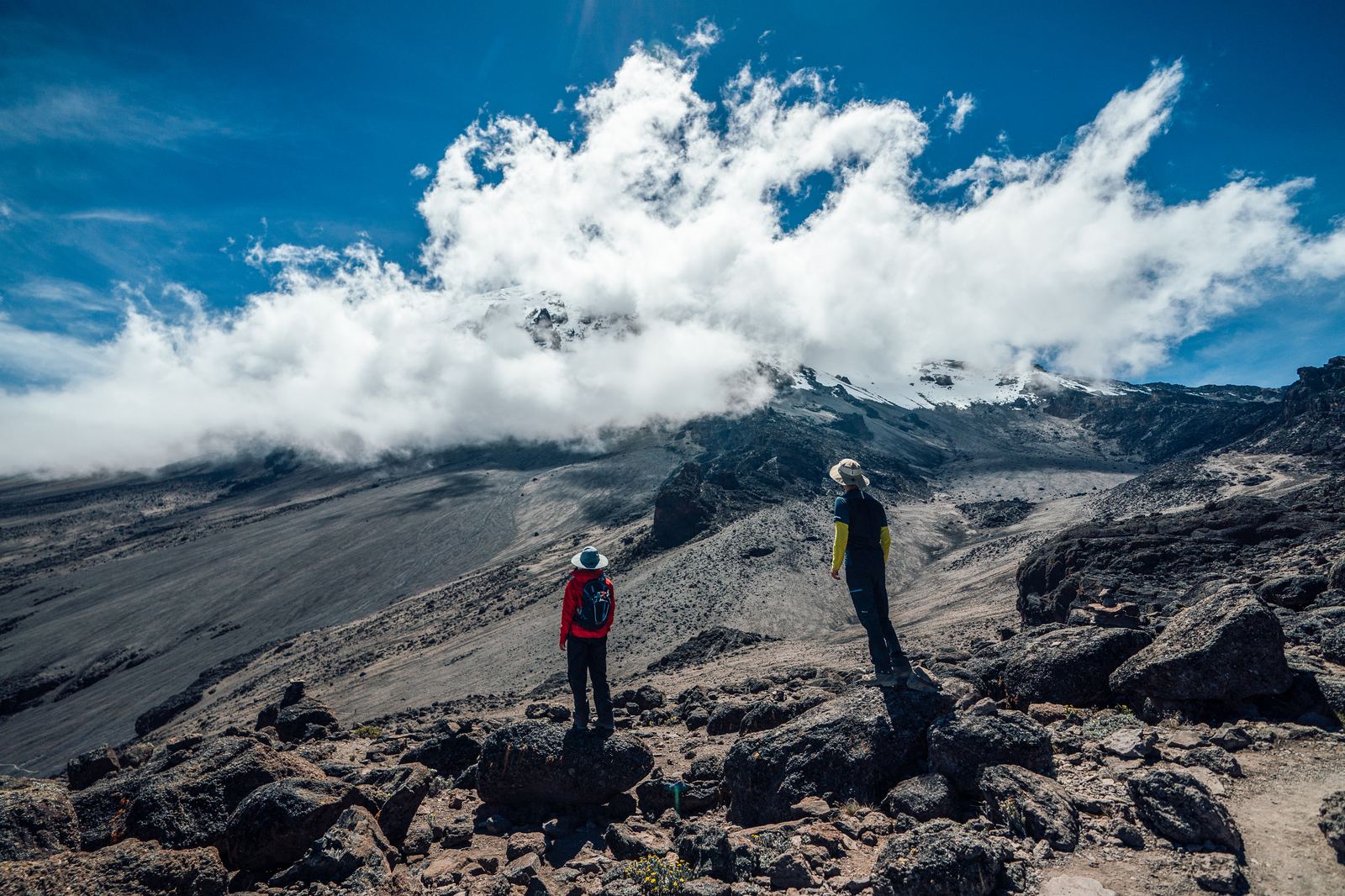 Two hikers standing on dark volcanic rock looking up at Kilimanjaro's summit half-hidden in cloud