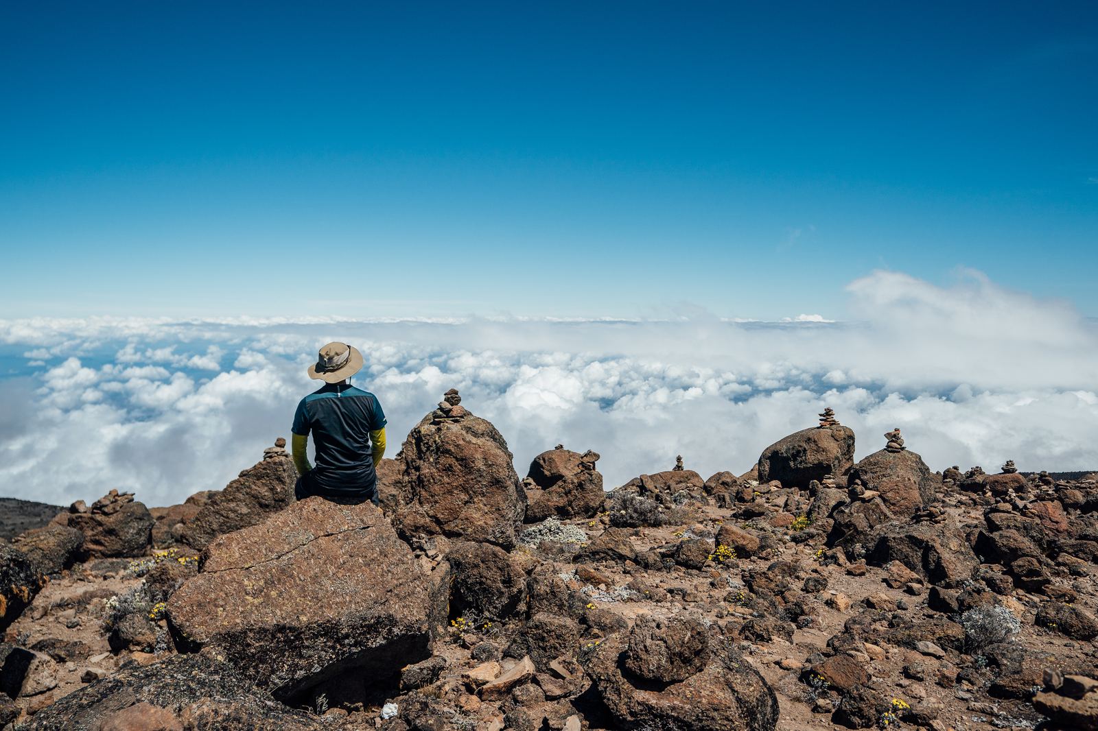 Hiker in a sun hat sitting on a rocky outcrop among stone cairns above a sea of clouds in bright daylight