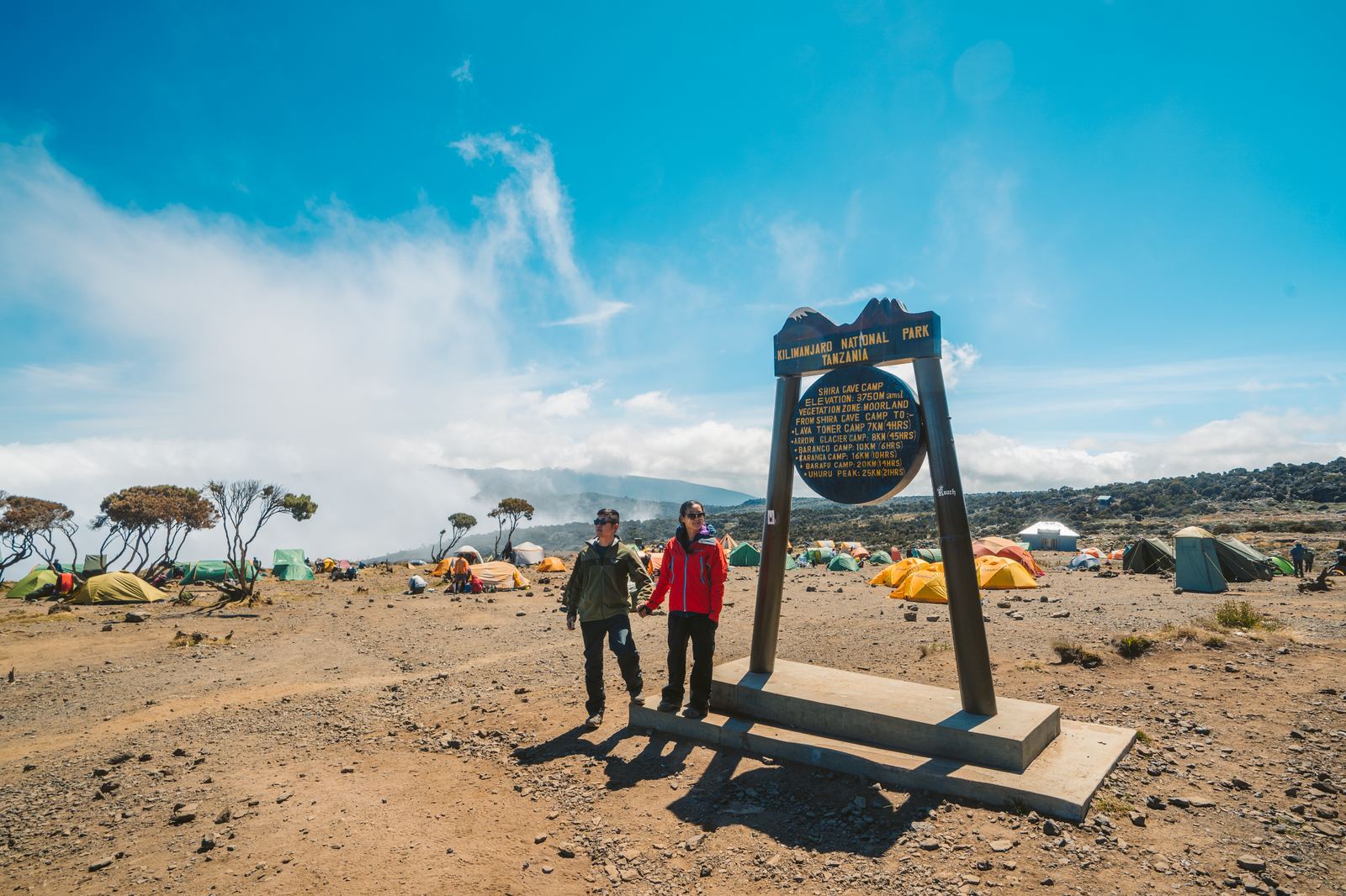 Jason and Grace at the Shira Cave Camp sign with expedition tents visible behind