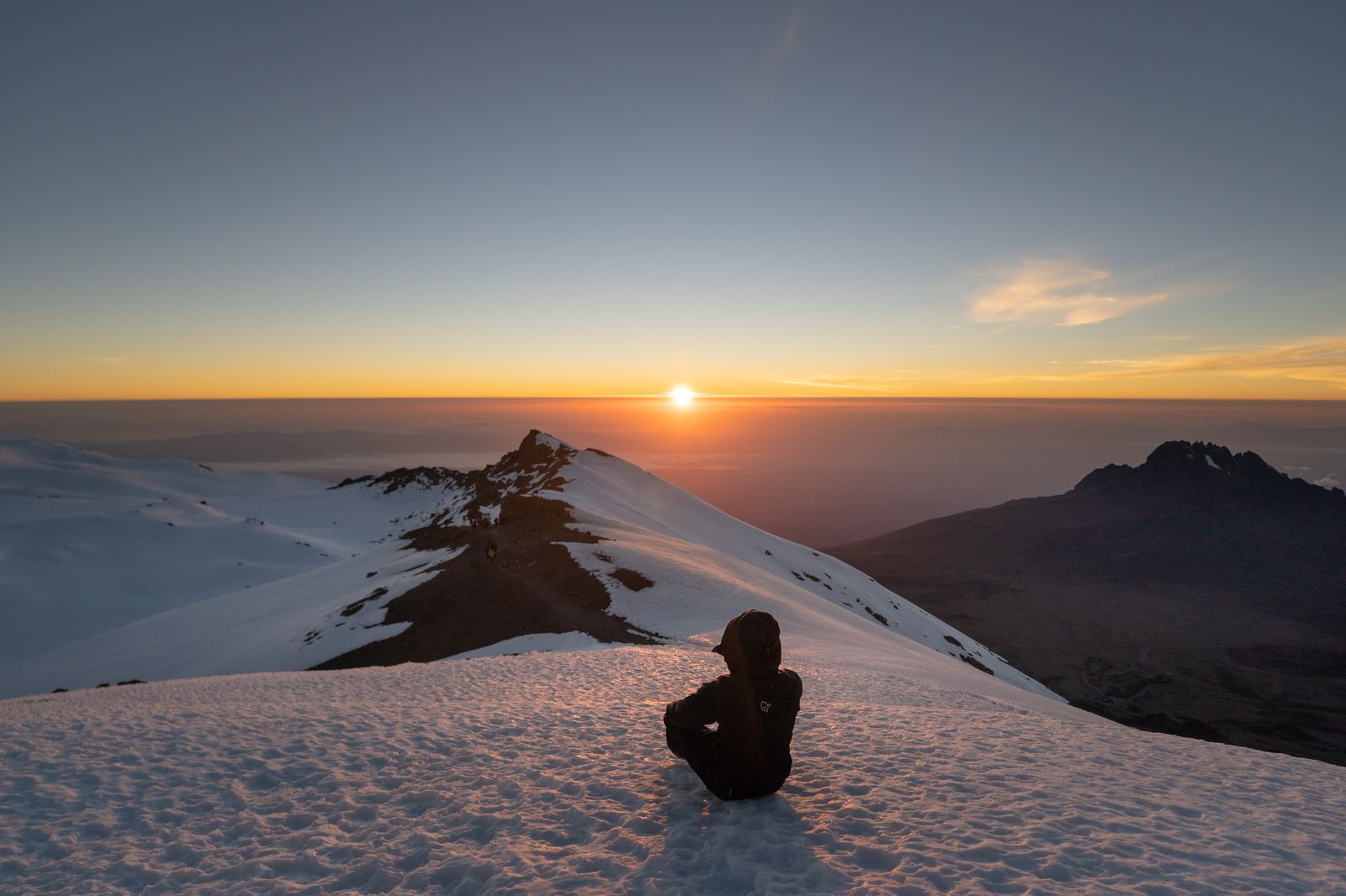 Climber sitting on the snowy crater rim of Kilimanjaro watching the sunrise over Mawenzi Peak