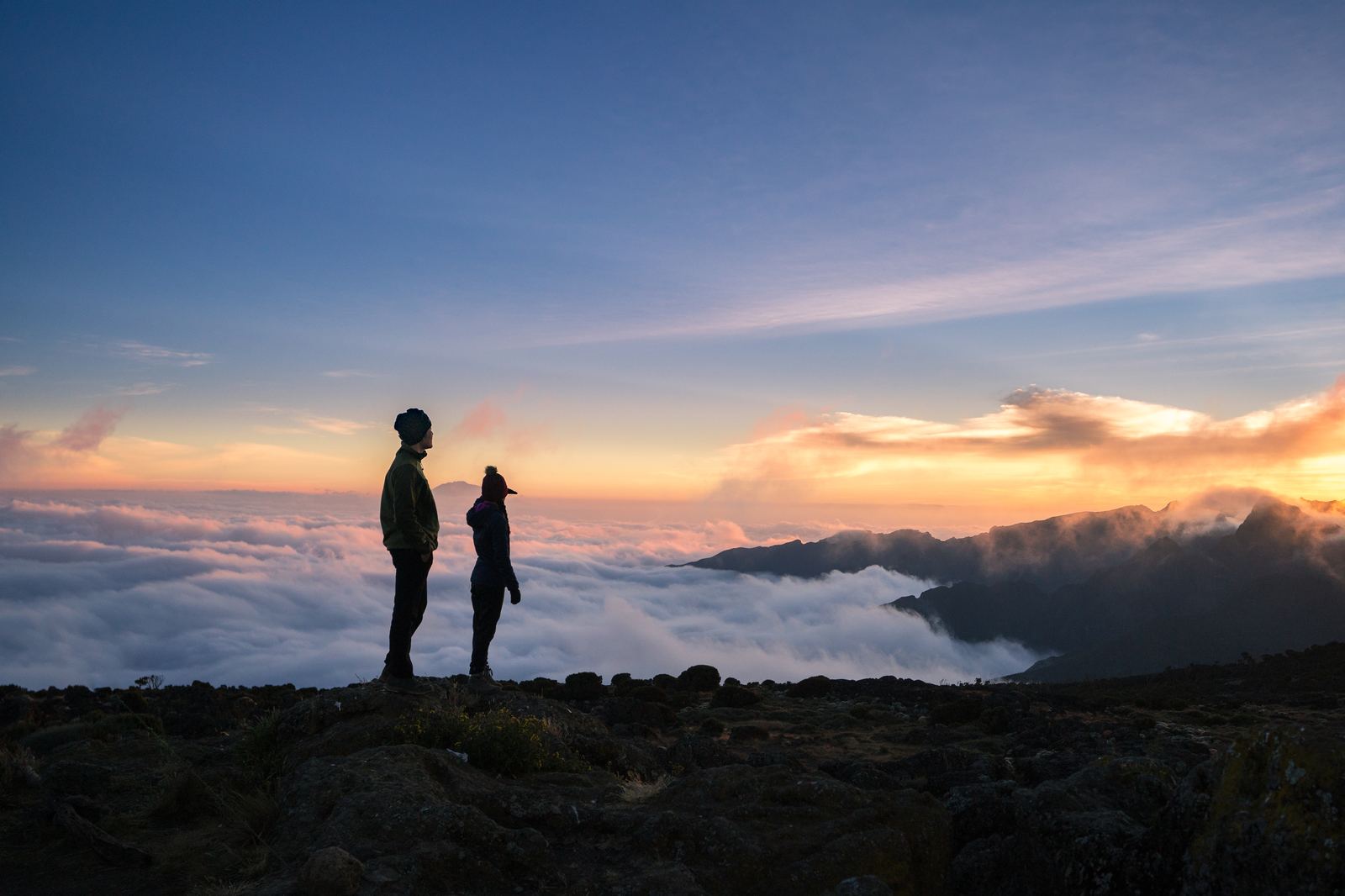 Two silhouetted figures standing above a sea of clouds at sunset on Kilimanjaro
