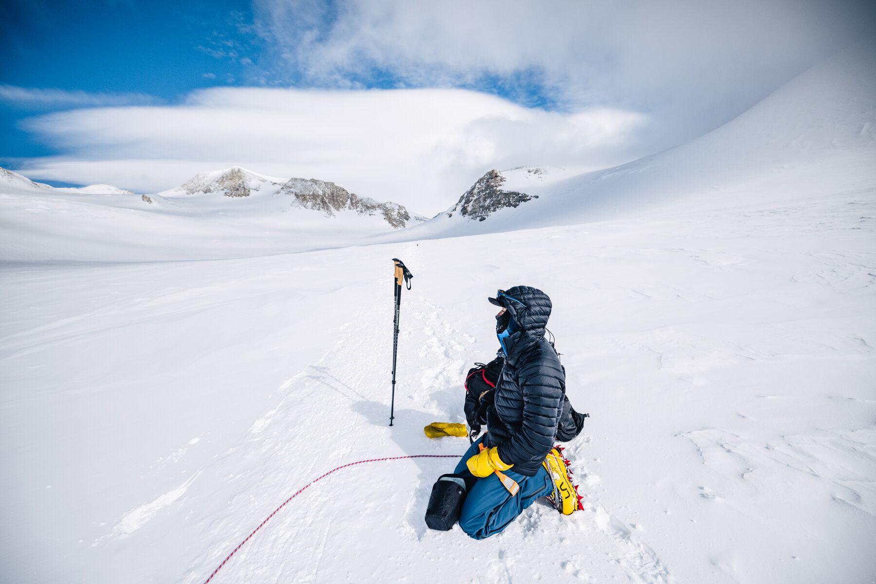 Climber kneeling alone in the snow on summit day, looking out at the rocky sub-peaks of the Vinson Massif under a clearing sky