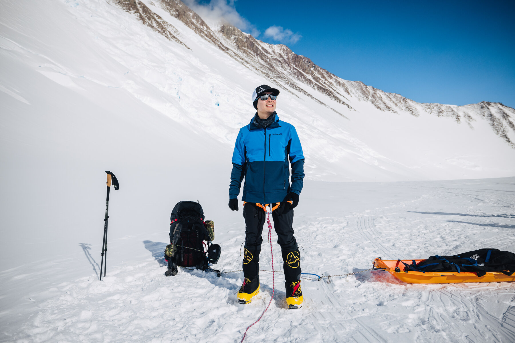 Climber in a blue softshell posing with poles and an orange haul sled at a rest stop on the Branscomb Glacier, the curving snow ridge behind