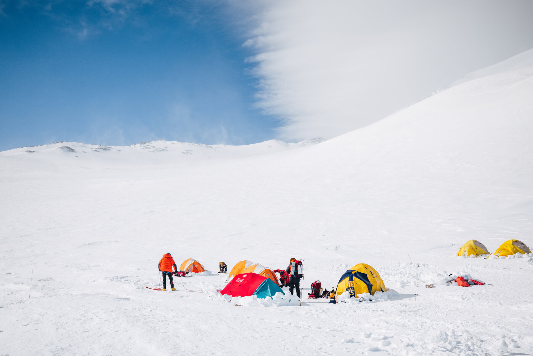 Cluster of colorful tents and climbers at camp on Vinson with a broad snow slope rising behind under blue sky