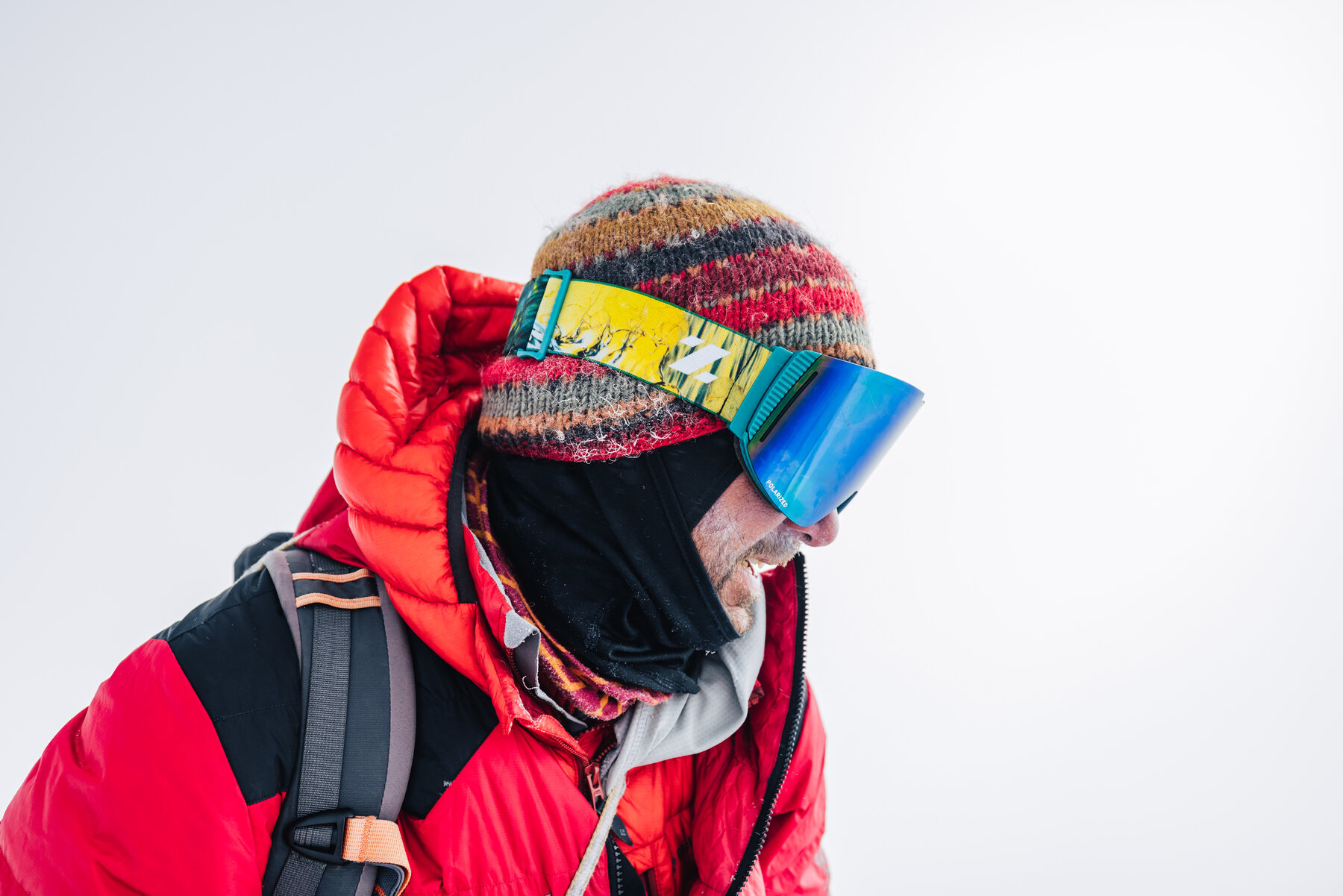 Tight portrait of a climber on the summit ridge — knit beanie, mirrored goggles, black face mask, red down jacket