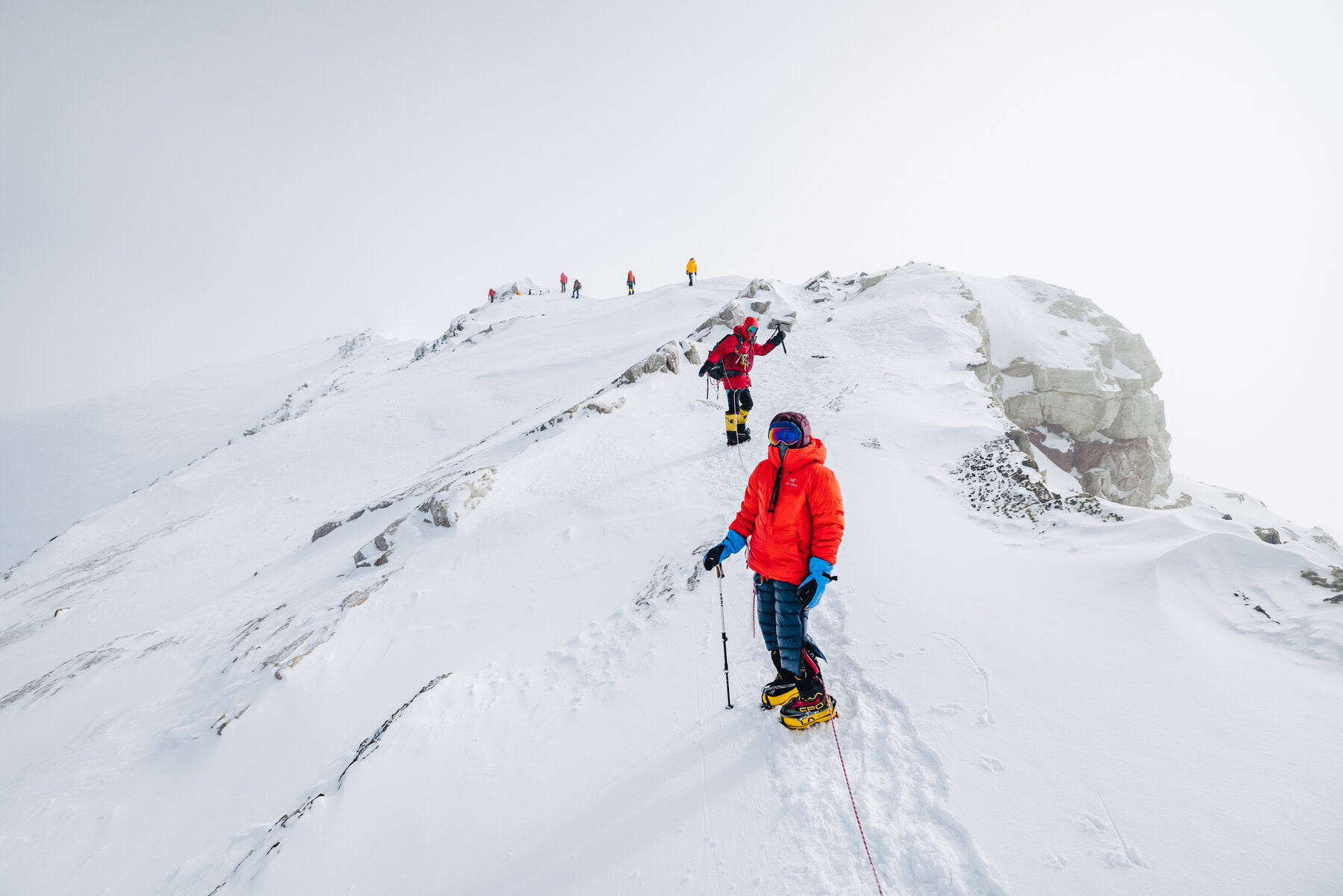 Pair of climbers in red and orange down jackets cresting the rocky Vinson summit ridge, more team members visible just ahead