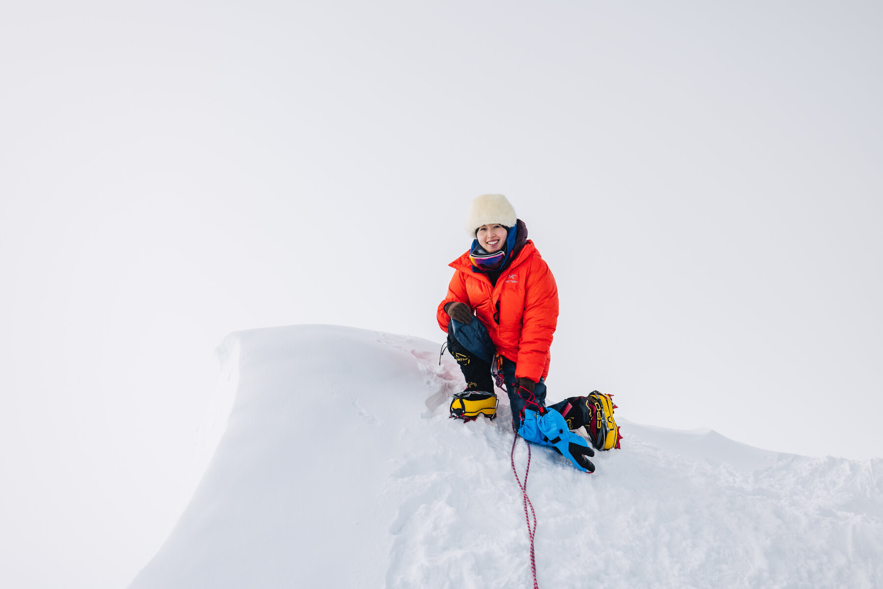 Grace in an orange puffy and white fur hat crouching on the corniced summit of Vinson with an ice axe