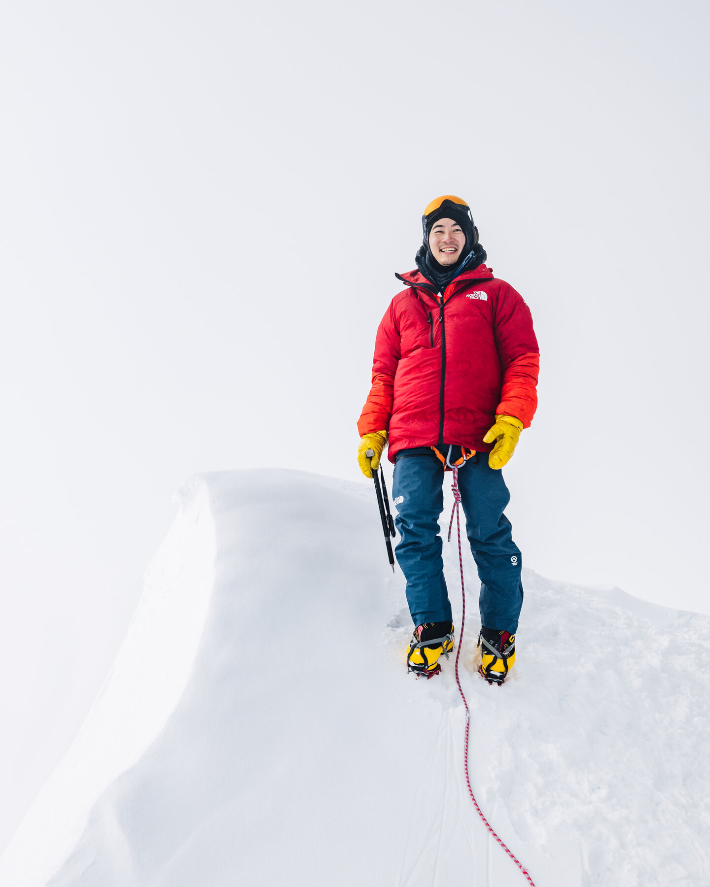 Climber in a red down jacket standing on the corniced summit of Vinson with the rope trailing down the snow