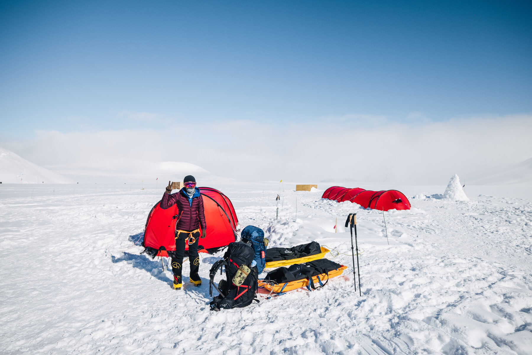 Two climbers with packs and loaded sleds beside red tents at camp on the glacier, one waving at the camera