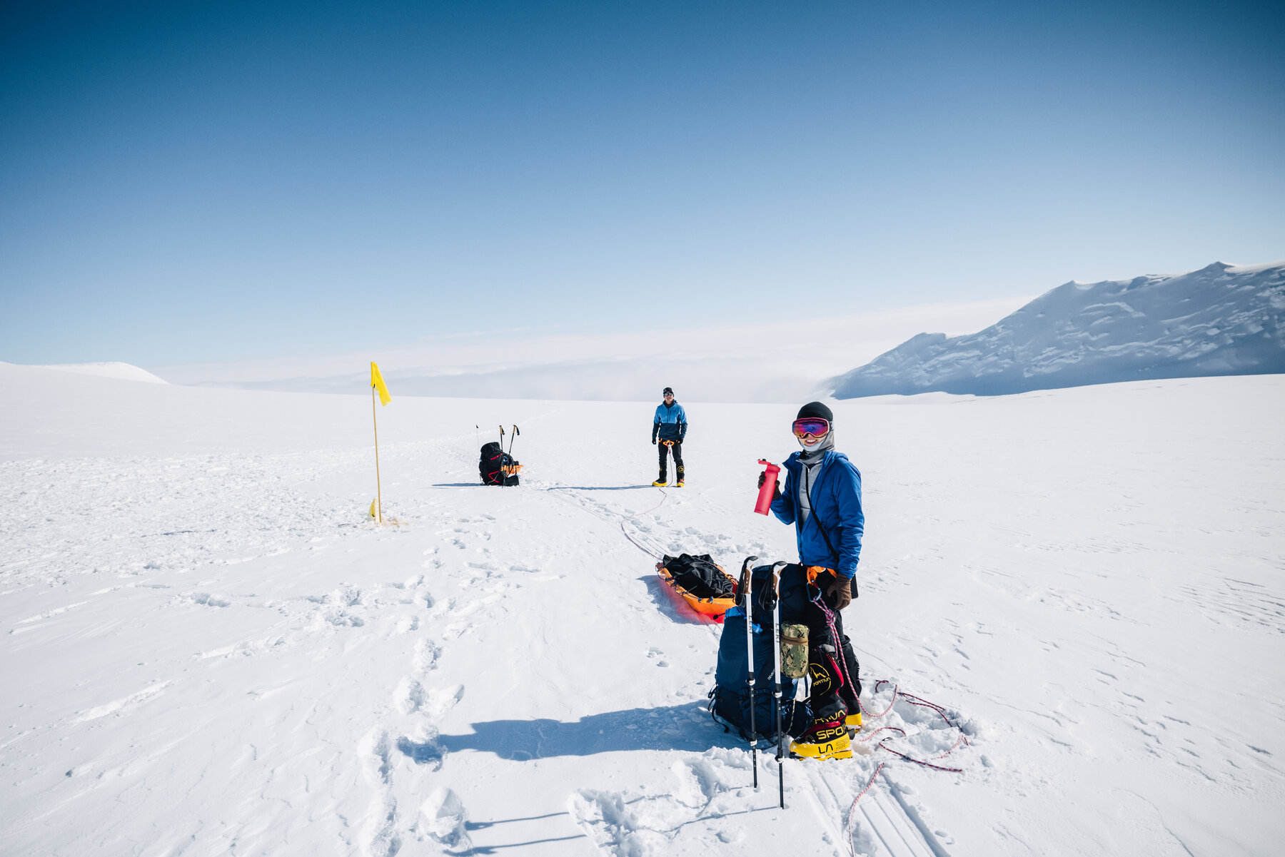 Two climbers with haul sleds pausing beside a marker wand on the glacier, snowy valley walls beyond