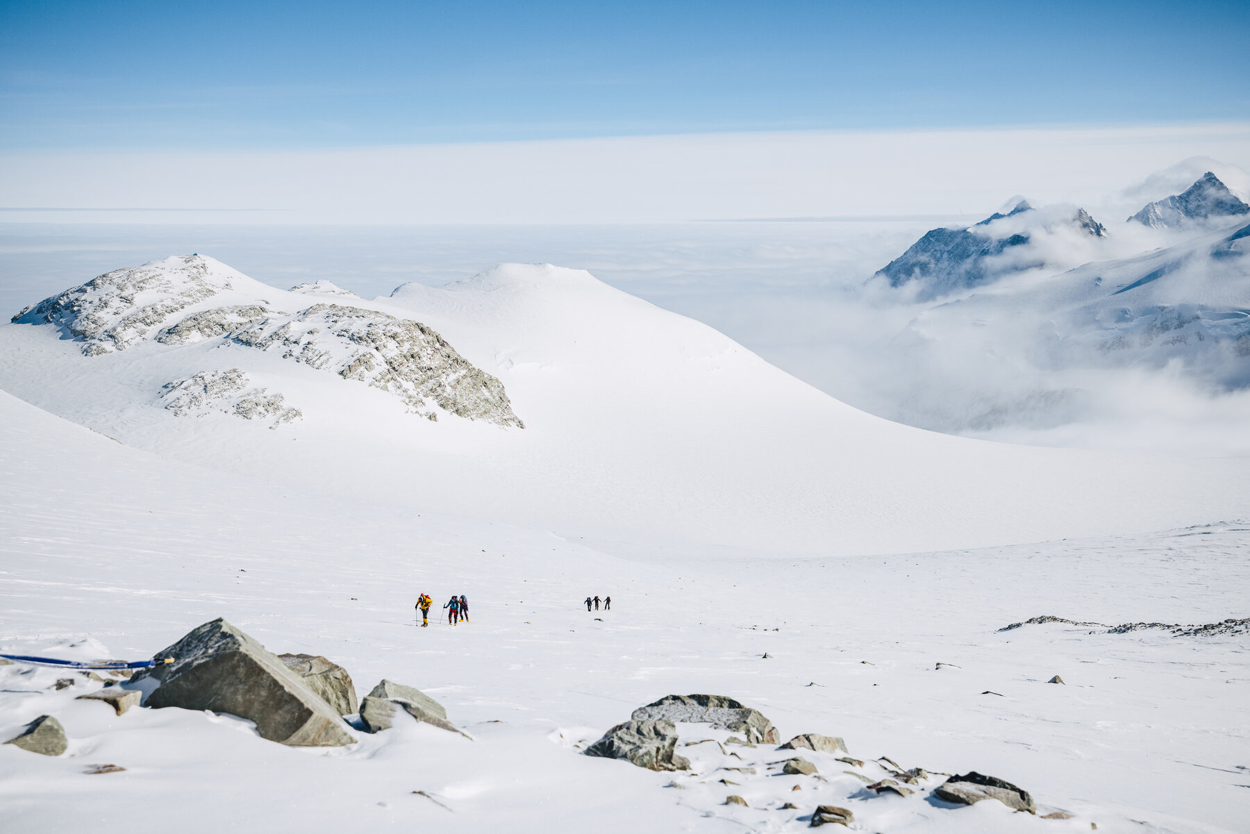 Wide view of climbers as small dots on a vast snowy landscape with rocky peaks and mountains behind
