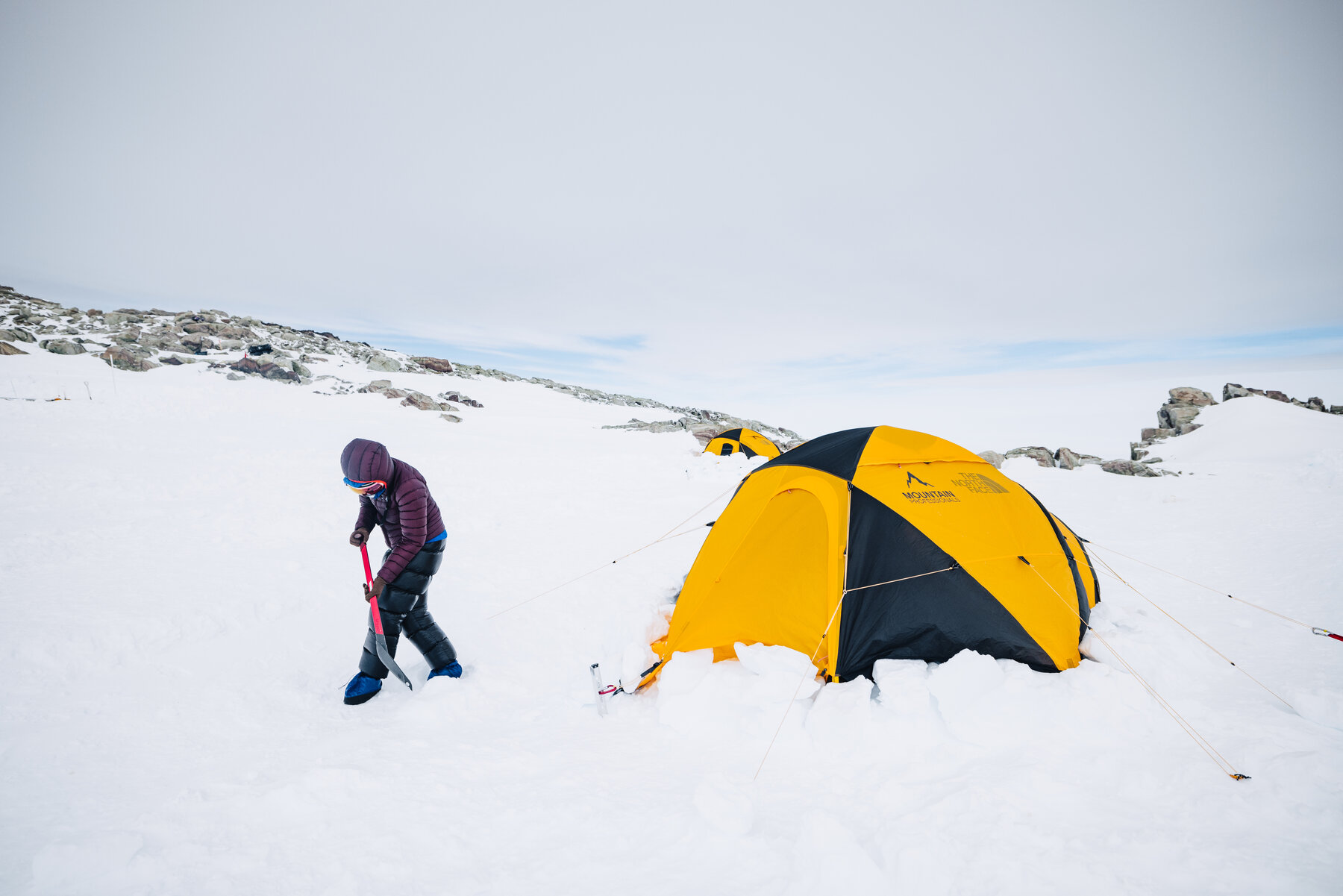 Climber shoveling snow around a yellow tent at camp on Vinson