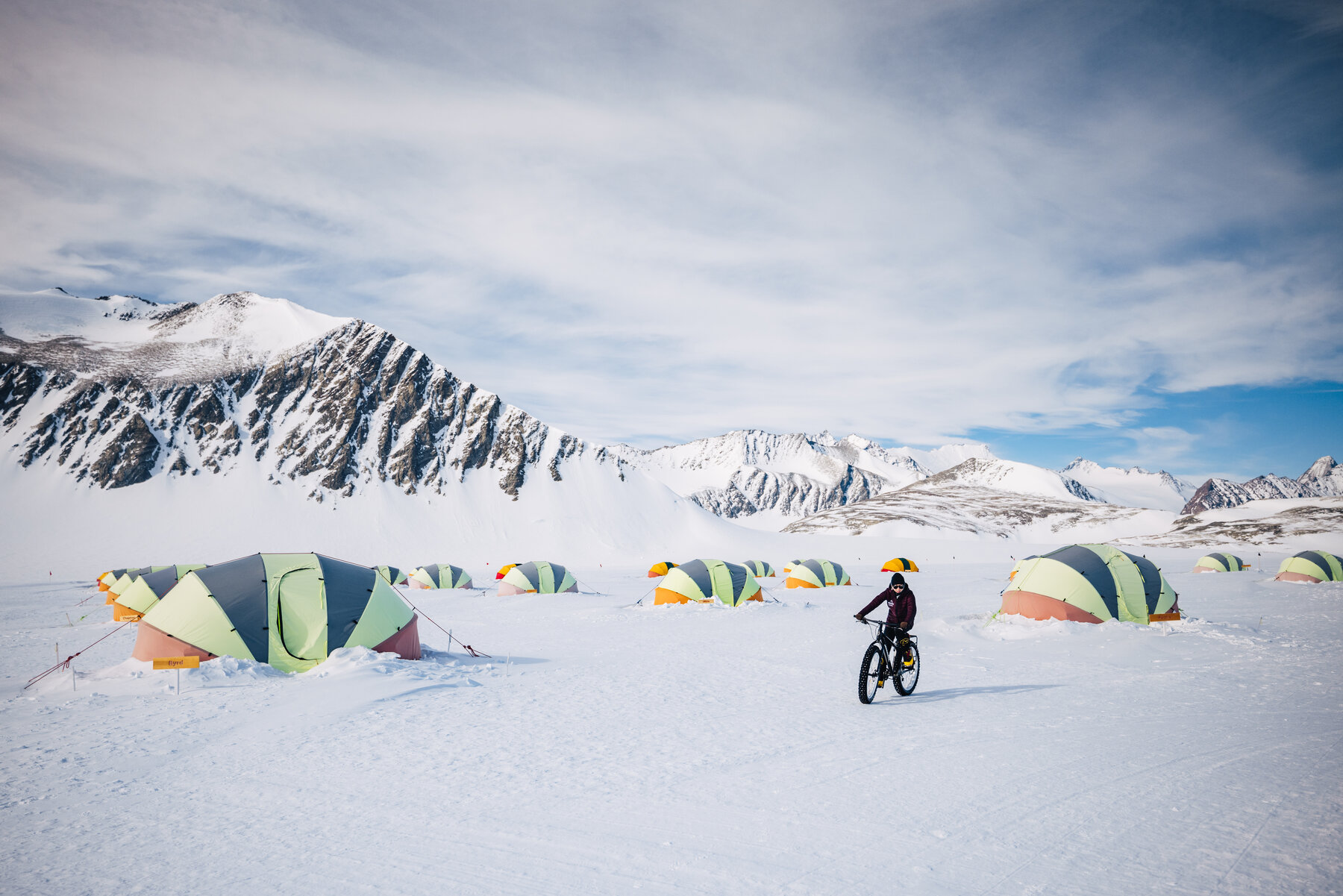 Cyclist on a fat tire bike crossing the Union Glacier tent village — colorful dome tents lined up against the snow-streaked Ellsworth peaks