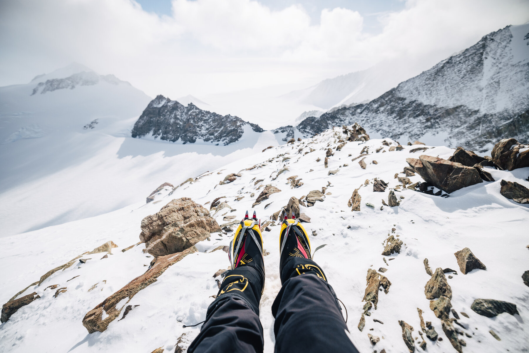 POV looking down past climbing boots and yellow gaiters from a rocky ridge above Vinson's glaciers, sharp peaks and clouds in the distance
