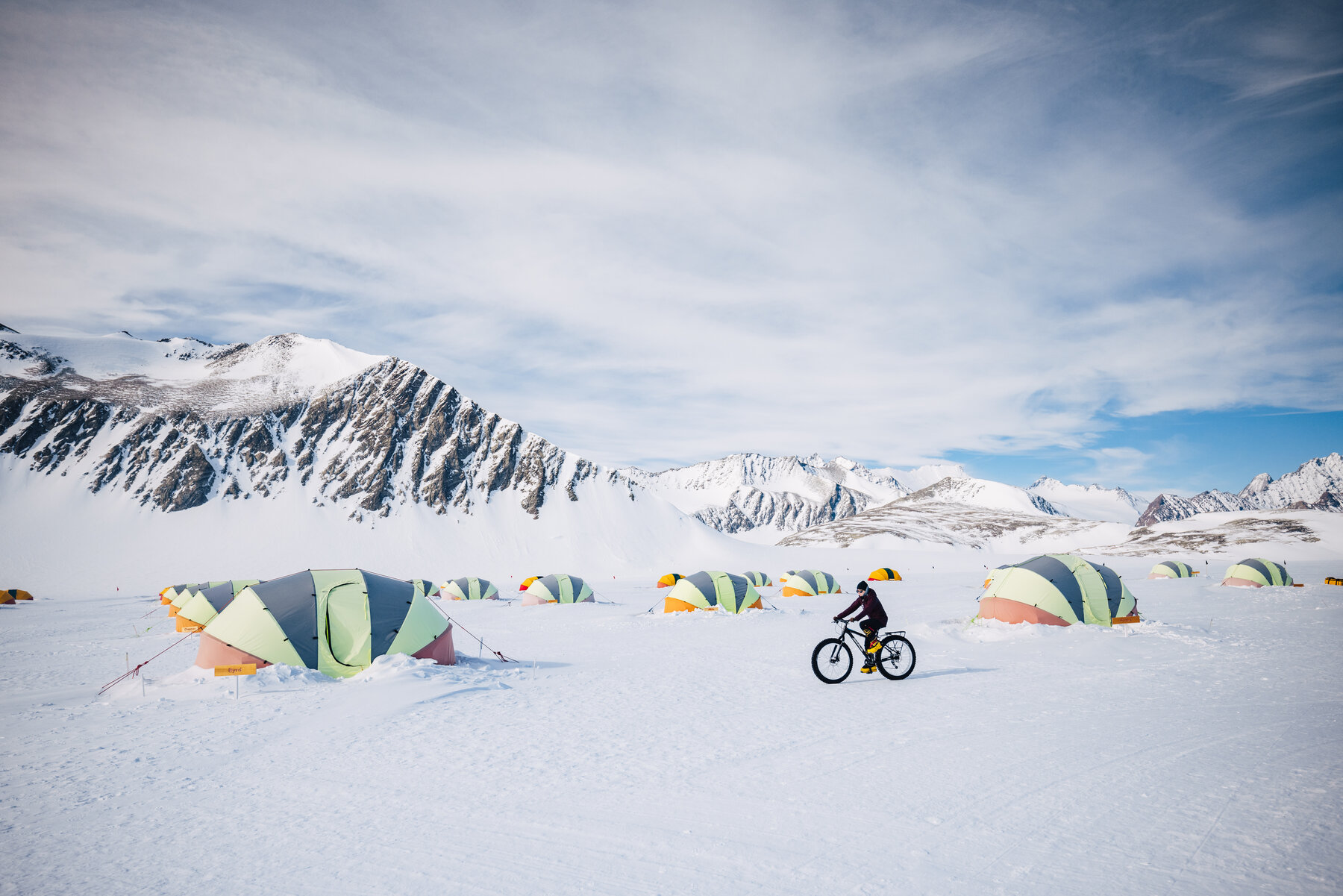 Cyclist on a fat tire bike passing the colorful dome tents at Union Glacier Camp with snowy peaks behind