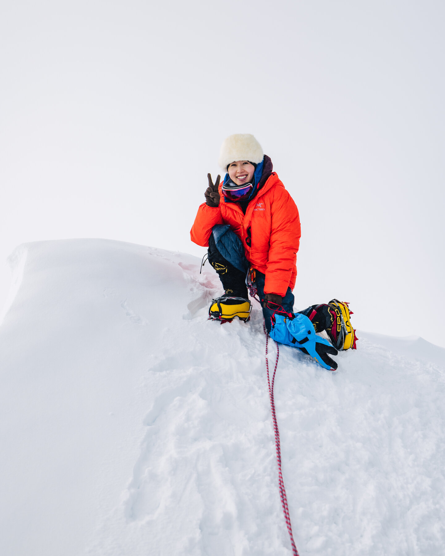 Grace in an orange puffy and white fur hat kneeling near the Vinson summit, flashing a peace sign through gloves