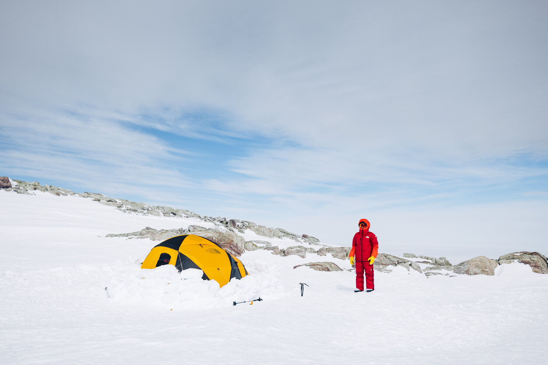 Climber in red suit standing next to a yellow tent at High Camp with rocky outcrops nearby